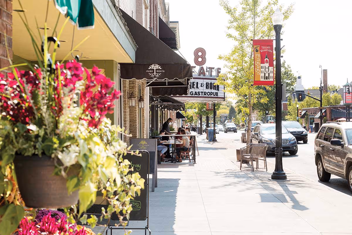 A sunny street scene with outdoor seating at a restaurant, colorful flowers in the foreground, parked cars along the street, and a lamppost with a red banner reading 'Uptown Westerville Engage'.