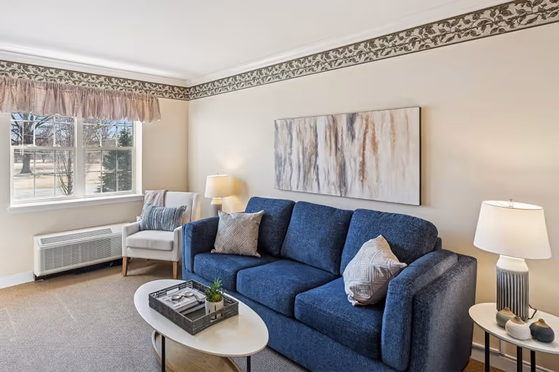 A cozy living room with a blue sofa adorned with two patterned pillows, a white armchair with a blue cushion, a white oval coffee table with a tray and small plant, two table lamps on either side, a large abstract painting on the wall, and a window with a pink valance letting in natural light.