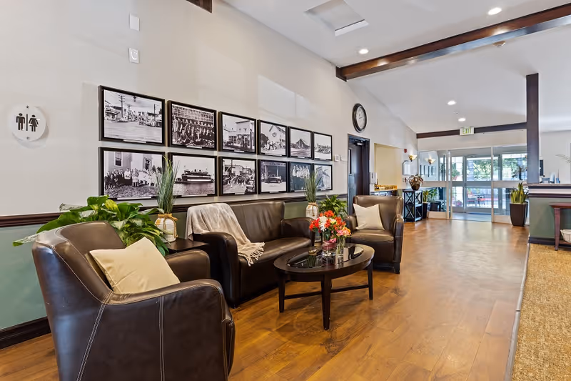 A bright and spacious seating area in a senior living facility with two brown leather armchairs and a matching sofa arranged around a dark wooden coffee table with a flower arrangement. The walls are decorated with a series of black and white framed photographs. There are plants placed near the seating area, and a clock is mounted on the wall above a door. The floor is wooden, and the space leads to a glass entrance door with an exit sign above it.