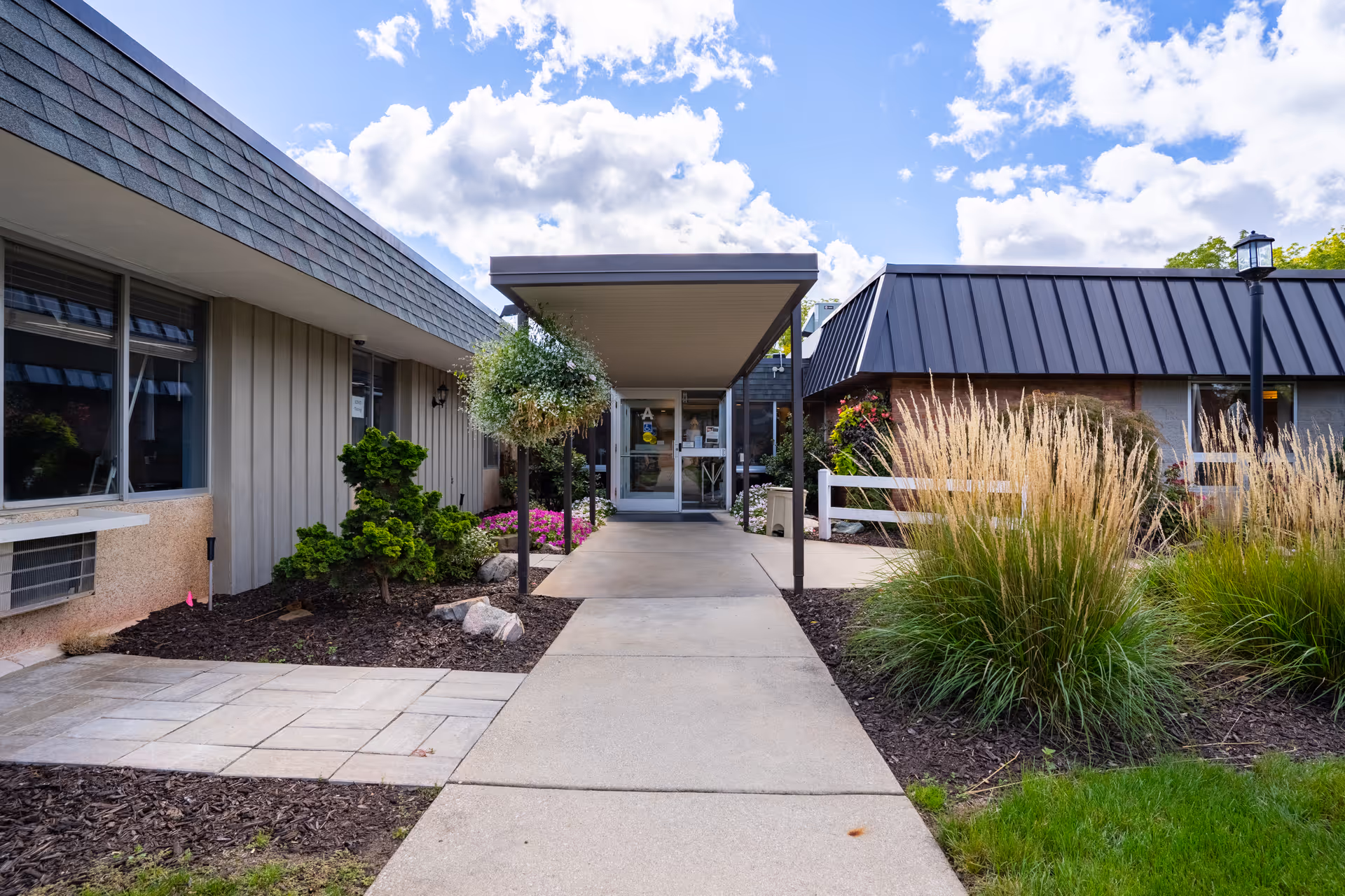 Entrance walkway to a single-story building with a covered entryway, surrounded by landscaped bushes, ornamental grasses, and flowers under a partly cloudy sky.
