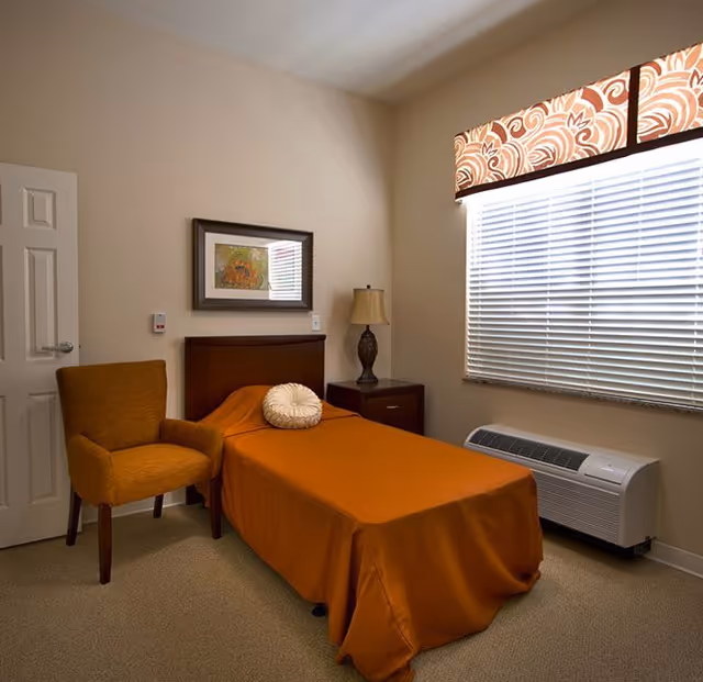 A small, neatly arranged bedroom with a single bed covered in an orange bedspread and a round decorative pillow. Next to the bed is a wooden nightstand with a lamp. There is an orange upholstered chair near the door, a framed picture on the wall, and a window with blinds and a patterned valance. Below the window is a white heating/cooling unit.