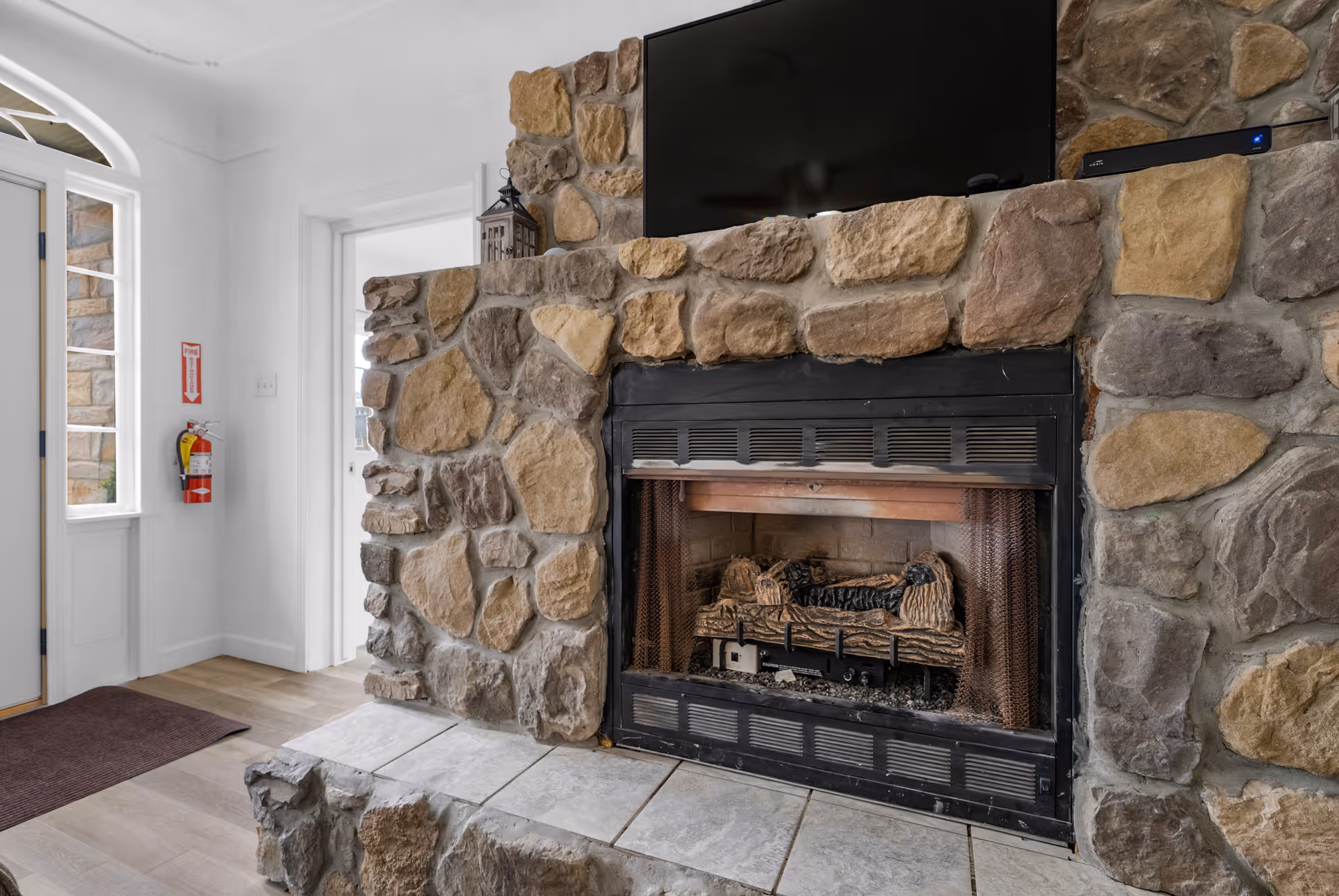 Interior view of a room featuring a stone fireplace with a mounted flat-screen TV above it. To the left, there is a door with glass panels and a fire extinguisher mounted on the wall next to it. The floor is wooden with a small rug near the door.