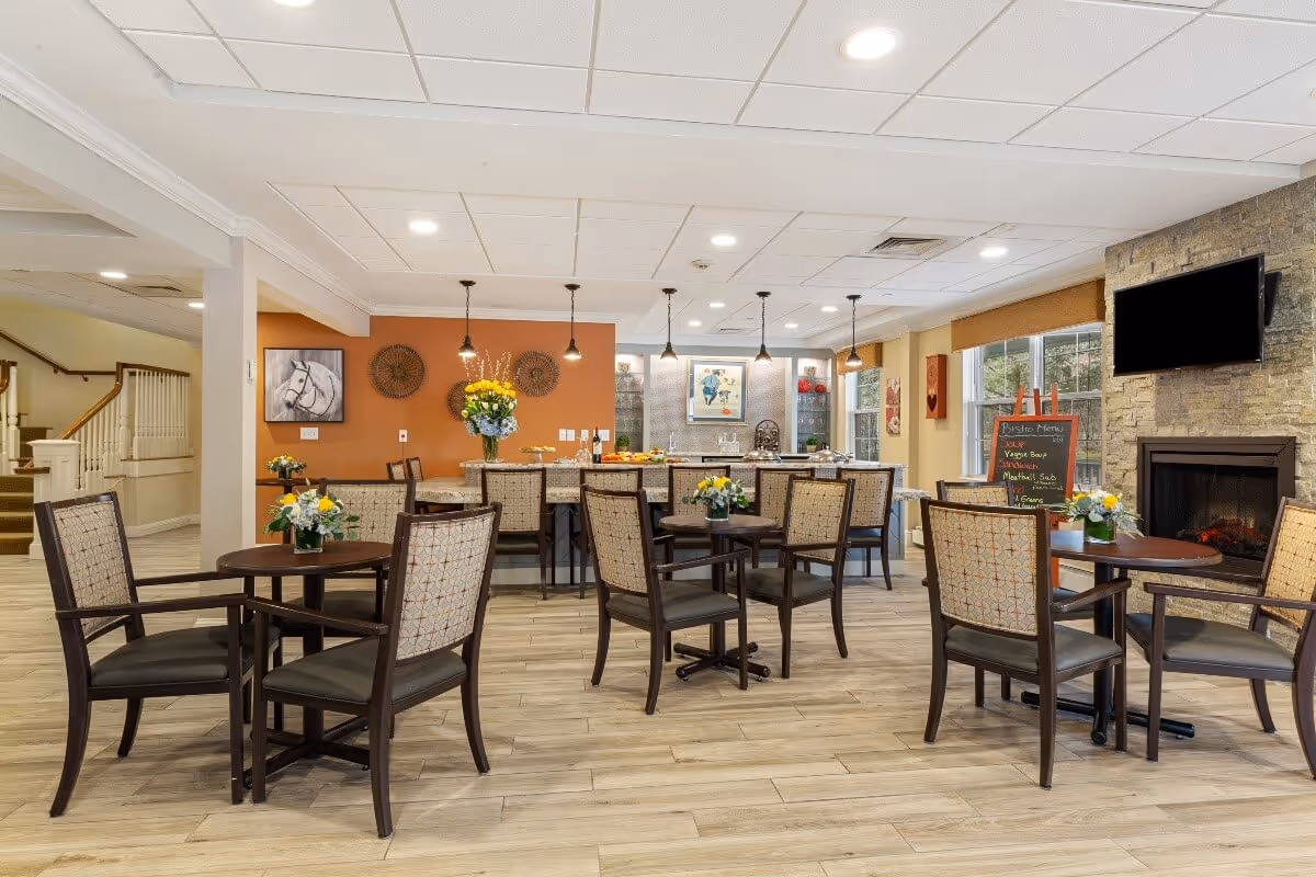 A bright and welcoming dining area in a senior living facility with multiple round tables and chairs, each table decorated with a small flower arrangement. In the background, there is a kitchen counter with bar stools, pendant lights hanging from the ceiling, and a chalkboard menu next to a stone fireplace with a mounted TV above it. The walls are painted in warm tones with decorative artwork and windows letting in natural light.