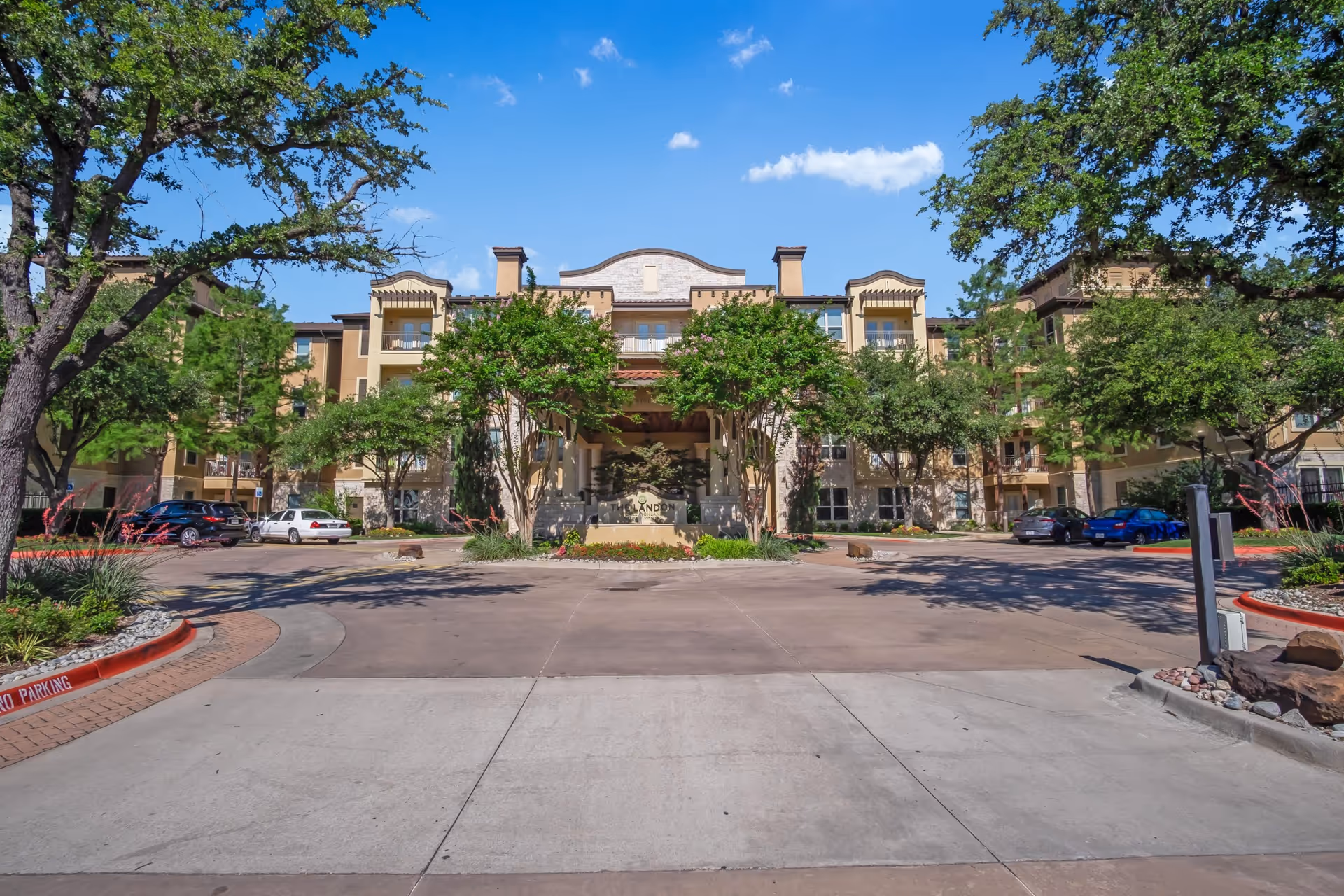 Front entrance of a multi-story senior living building with a circular driveway, landscaped trees, and parked cars under a blue sky.