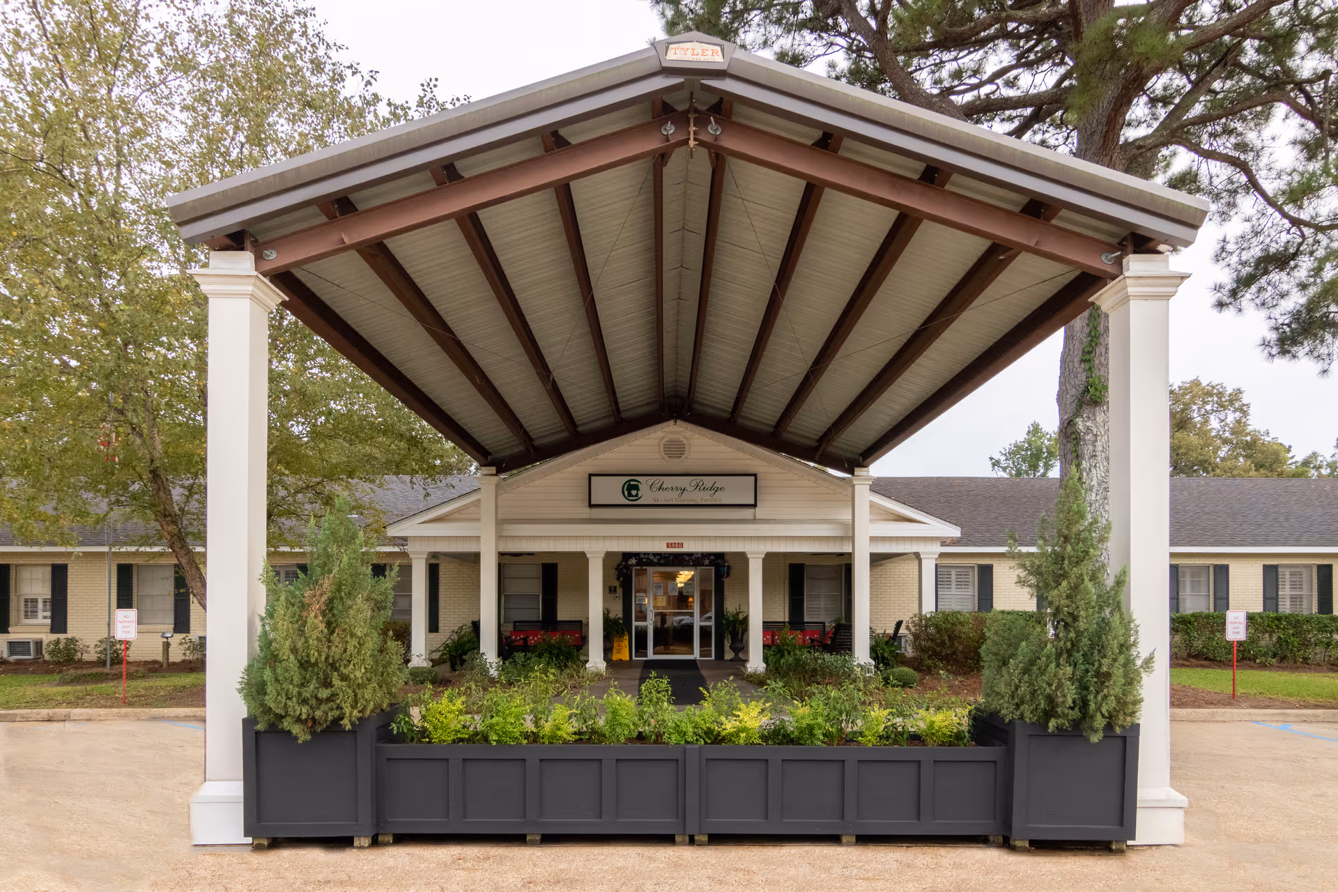 Front exterior view of Cherry Ridge Skilled Nursing Facility showing a covered entrance with white pillars, a garden bed with green plants, and a sign above the entrance displaying the facility's name.