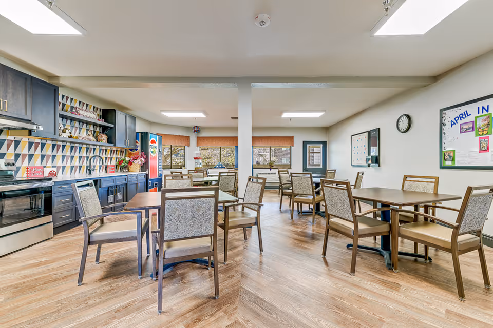 A bright and clean dining area in a senior living facility with multiple wooden tables and cushioned chairs. The room features a kitchen area with dark cabinets, a stove, and a colorful geometric backsplash. There are windows with brown blinds, a Pepsi vending machine, a popcorn machine, and bulletin boards on the walls.