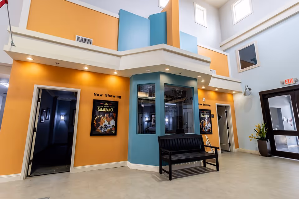 Interior view of a senior living facility lobby area with high ceilings and large windows. The walls are painted in warm orange and light blue tones. There is a black bench in front of a blue ticket booth or reception window. On the orange walls, there are movie posters for 'Casablanca' with 'Now Showing' signs above them. To the right, there is a glass exit door with a potted plant nearby.