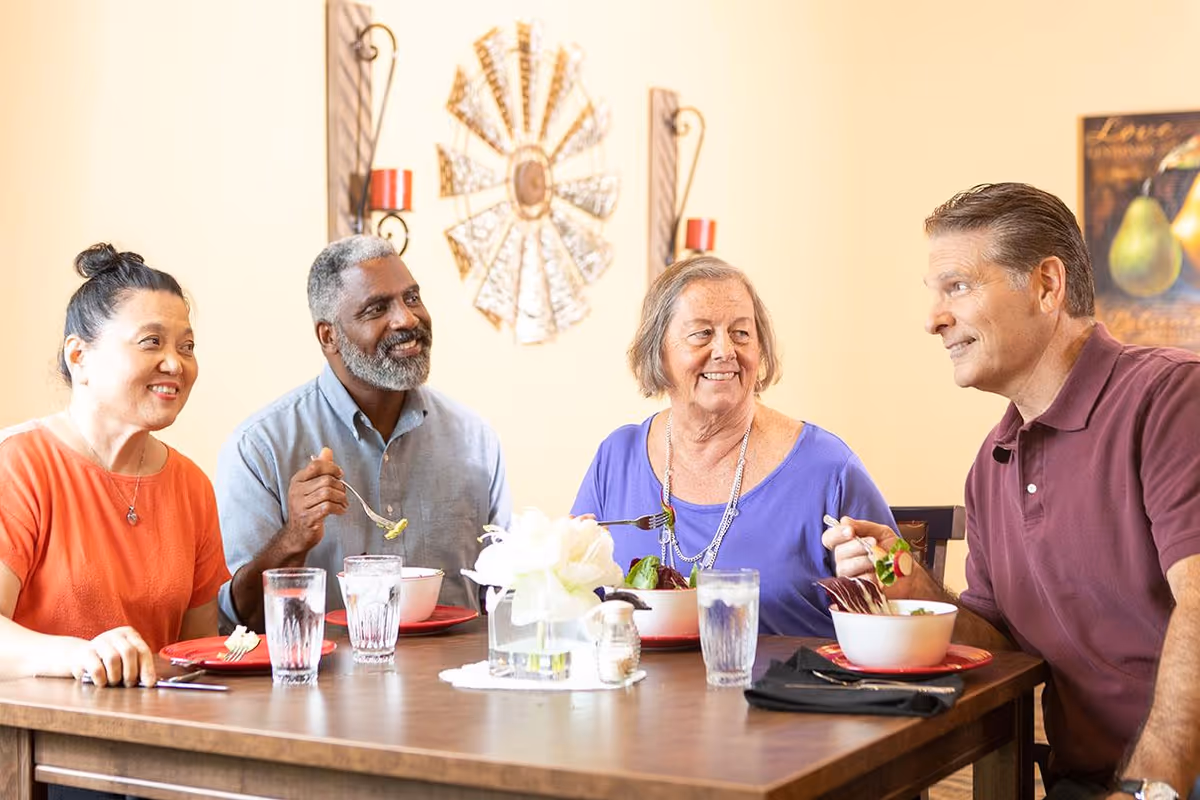 Four adults sitting around a dining table enjoying a meal together, smiling and engaging in conversation. The table is set with bowls of food, glasses of water, and a centerpiece with white flowers. The background features wall decorations including a metal windmill art piece and a framed picture of pears.