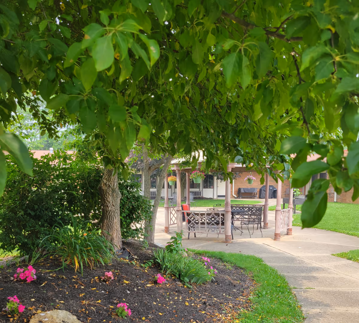 A shaded outdoor garden area with a tree and flower bed in the foreground. A gazebo with metal chairs and tables is visible on a concrete patio, surrounded by green grass and hanging flower pots. The background shows part of a brick building with windows and a covered porch.