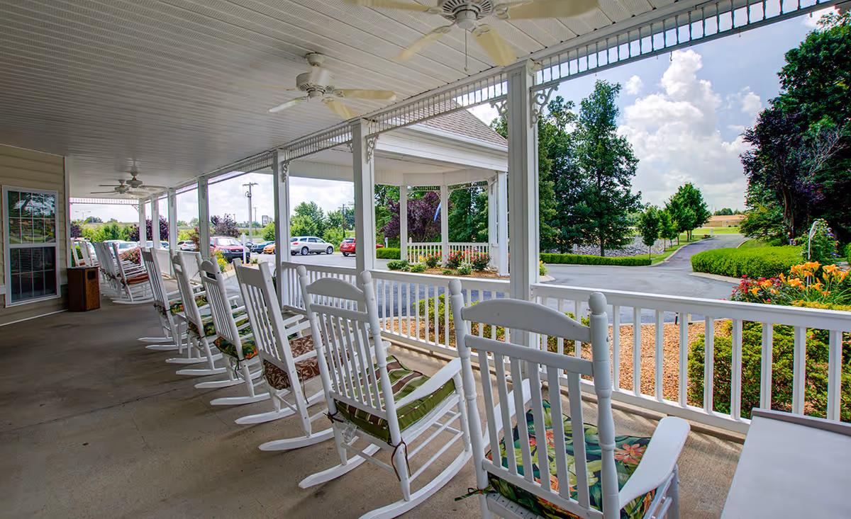 Covered front porch with a row of white rocking chairs overlooking landscaped grounds and a driveway.