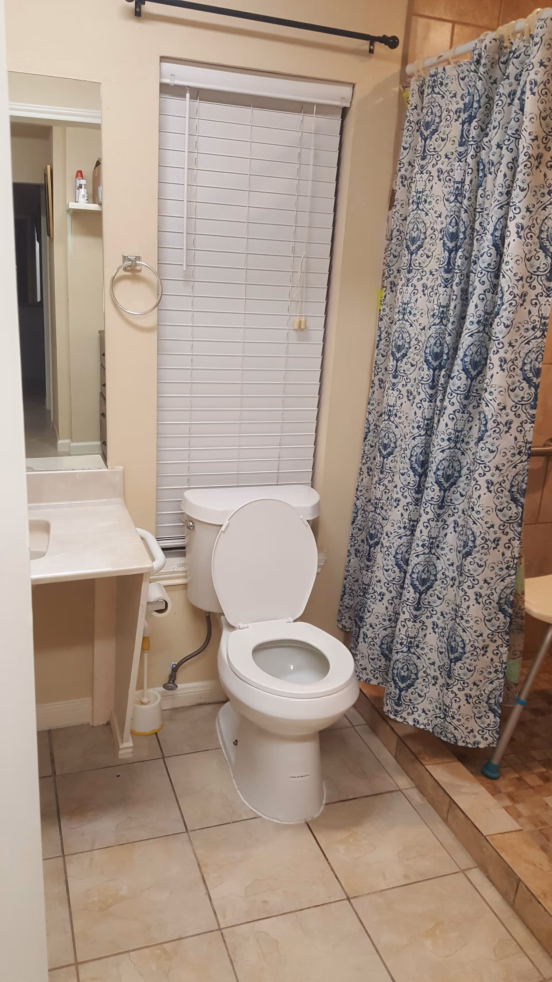 Small bathroom with a toilet, a sink, a window with closed blinds, and a patterned shower curtain over a tiled floor.