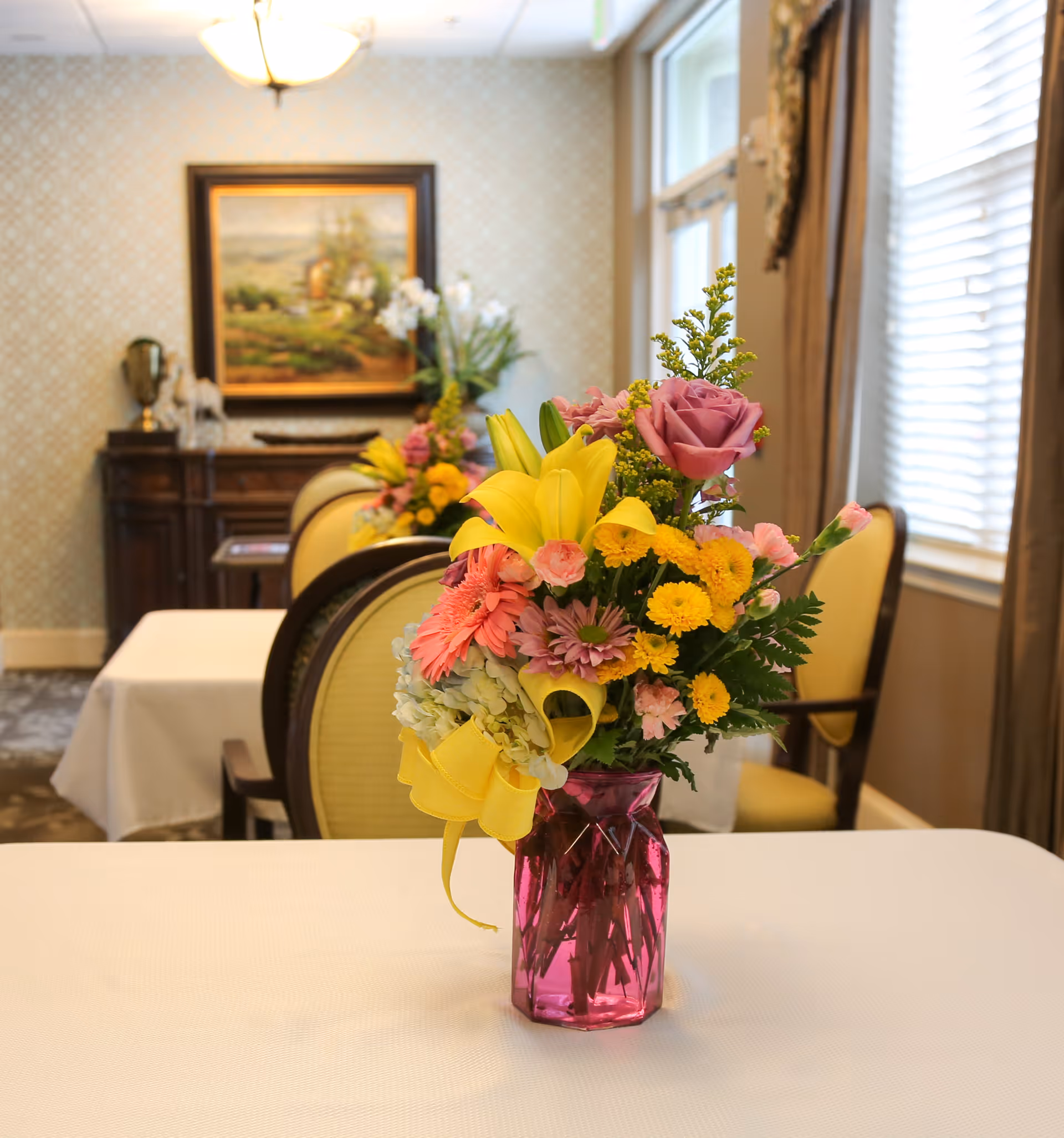 A colorful bouquet in a pink glass vase sits on a table in a bright dining room with upholstered chairs and a framed painting in the background.