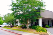 Exterior view of a building entrance with a covered driveway, surrounded by green trees and shrubs, with a white vehicle parked on the left side.