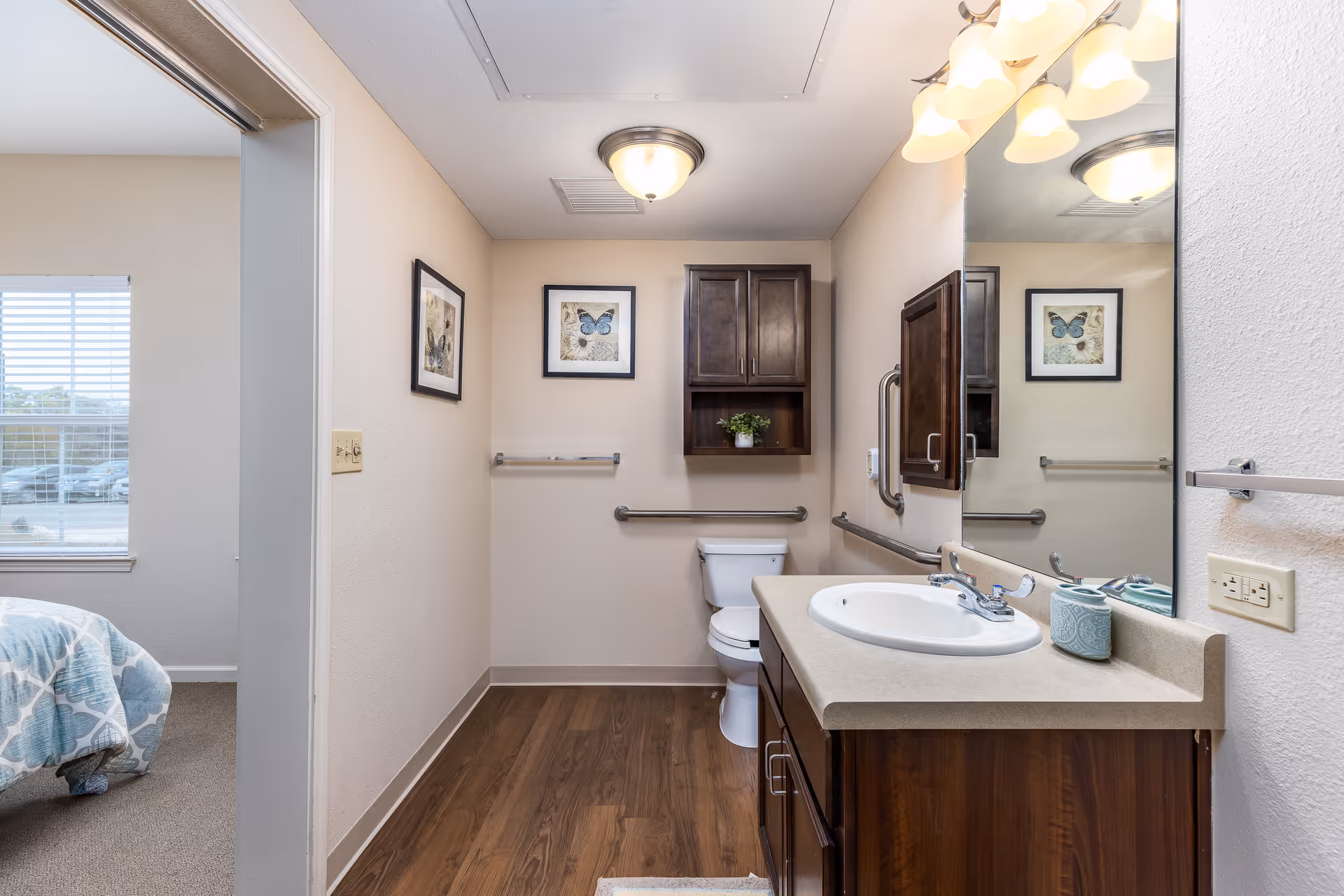 Accessible bathroom with a sink and vanity, toilet, wall cabinets, grab bars, and a large mirror, with a bedroom visible through the doorway.