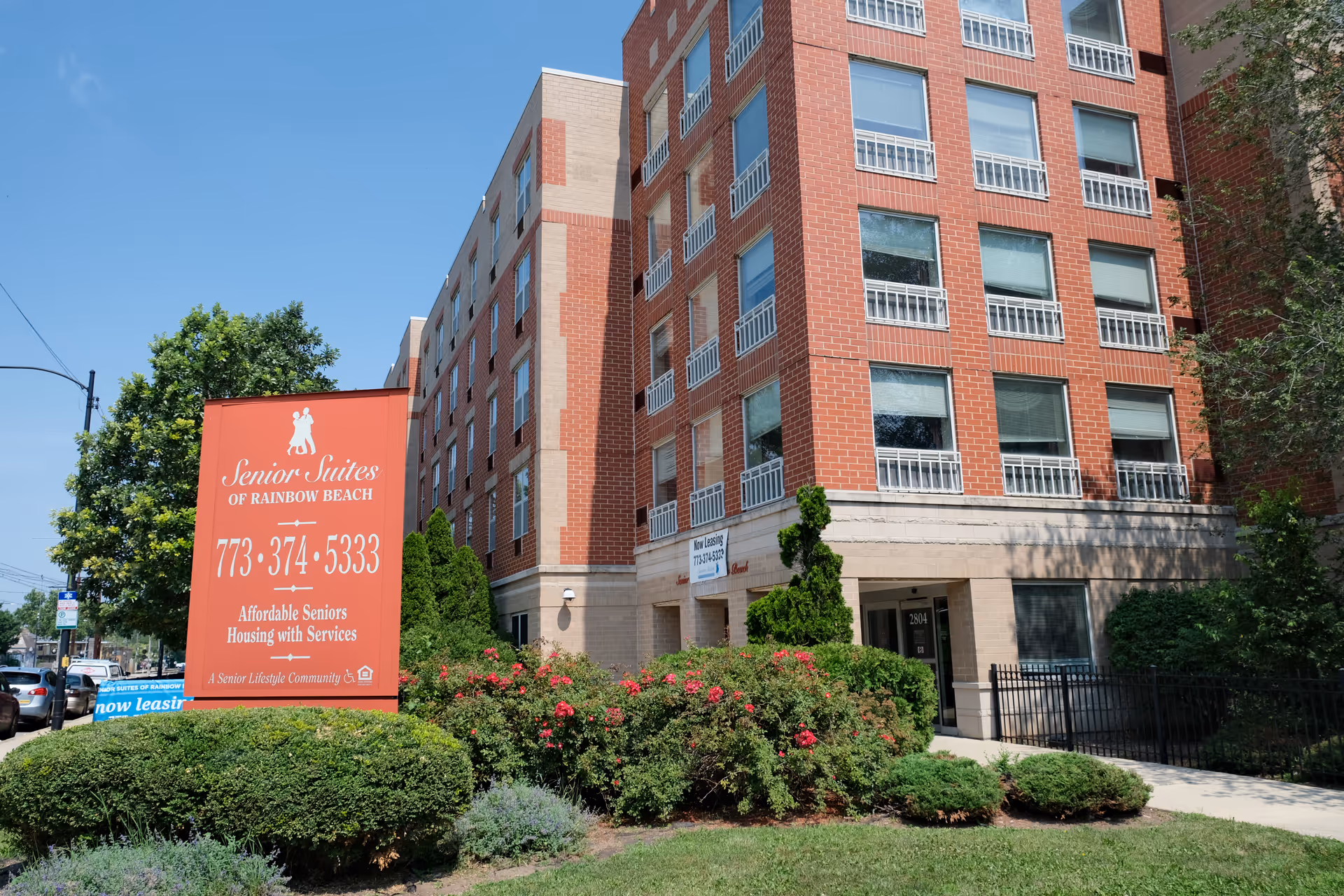 Exterior view of a multi-story brick building with large windows and a landscaped garden in front. A red sign in the foreground reads 'Senior Suites of Rainbow Beach, 773-374-5333, Affordable Seniors Housing with Services, A Senior Lifestyle Community'.