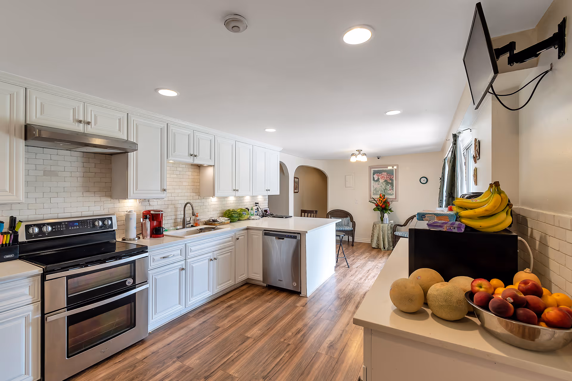 A bright and spacious kitchen with white cabinetry, stainless steel appliances including a stove and dishwasher, and a white countertop island. The kitchen has wooden flooring and a tiled backsplash. On the right side countertop, there is a bowl of fresh fruit including bananas, cantaloupes, and peaches. In the background, there is a small seating area with two chairs, a small table with a flower vase, and a wall-mounted TV on the right.
