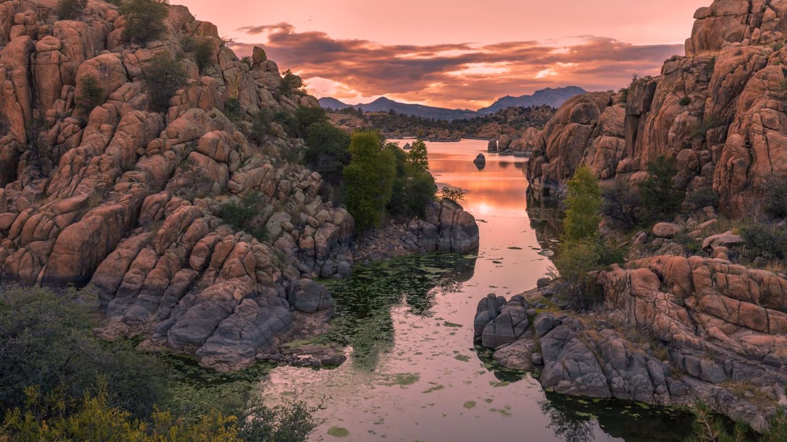 A scenic view of a rocky canyon with a narrow river or lake running through it, surrounded by large rock formations and sparse trees under a colorful sunset sky.