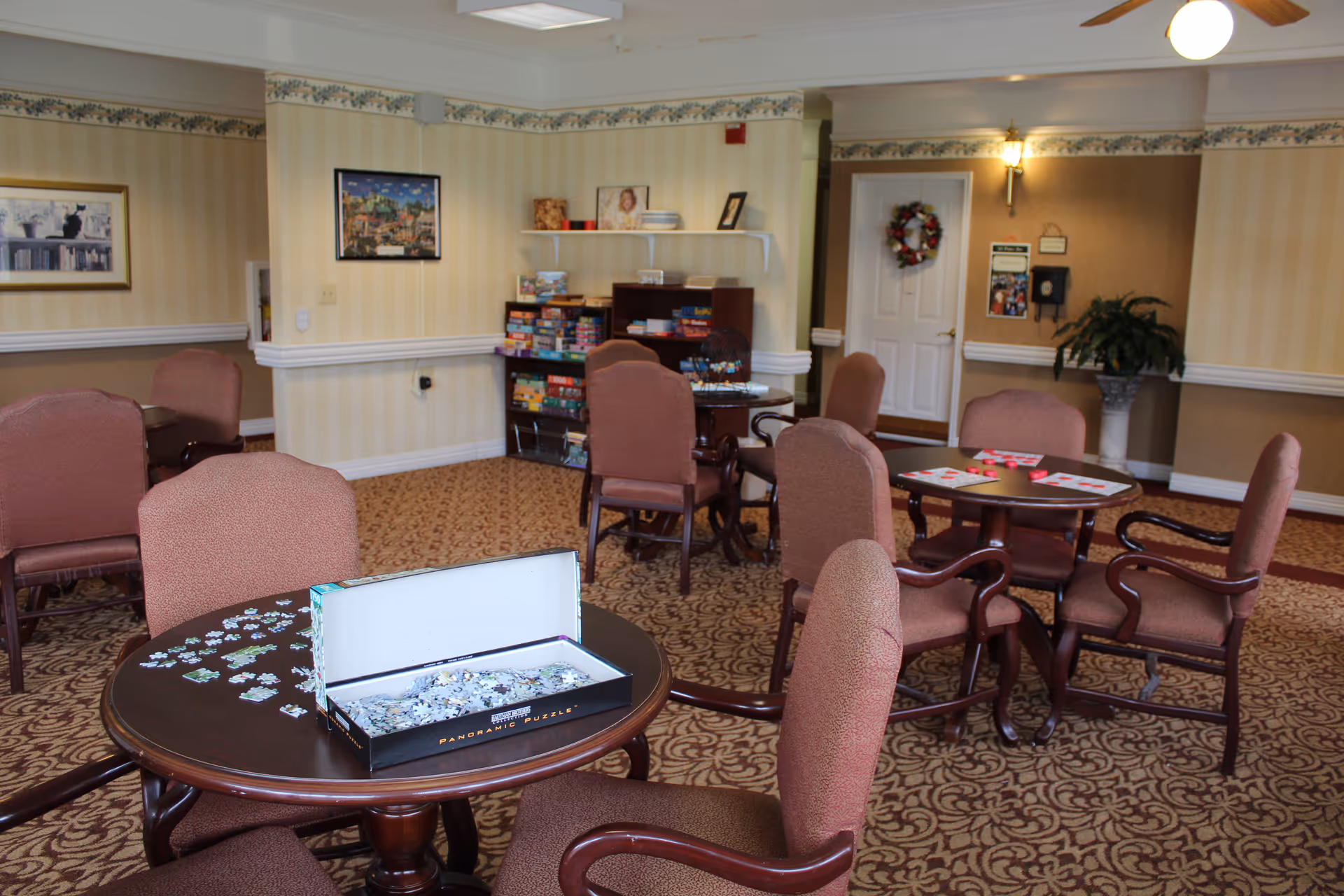 A cozy activity room with several round tables and cushioned chairs arranged around them. One table has a partially completed jigsaw puzzle with the puzzle box open. Another table has playing cards spread out. Shelves in the background hold board games and puzzles. The room has patterned carpet, wallpaper with a floral border, framed pictures on the walls, and a door decorated with a wreath.
