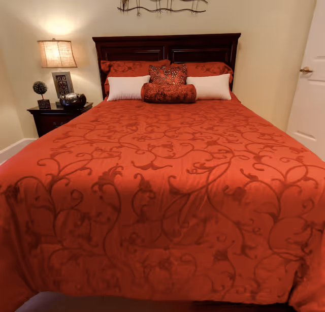 A neatly made bed with a dark wooden headboard, covered with a red bedspread featuring a floral pattern. The bed has multiple pillows in red and white. To the left of the bed is a nightstand with a lamp, a small decorative plant, and a silver decorative object. The walls are light-colored, and there is a closed white door visible on the right side.