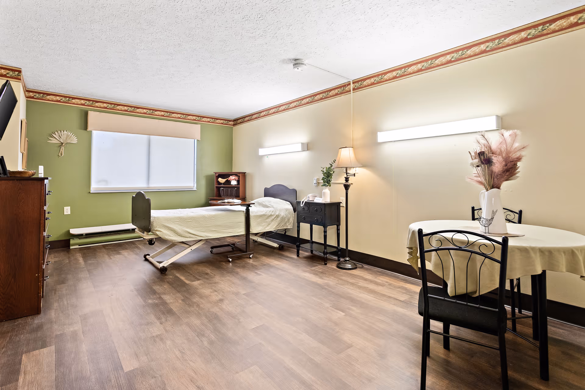 A nursing center bedroom with a single hospital bed, a small wooden dresser, a nightstand with a lamp and phone, a round table with two chairs and a vase with dried flowers, and a large window with a white blind. The walls are painted green and beige with a decorative border near the ceiling, and the floor is wood.