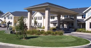 Exterior view of Mulberry Gardens Senior Living facility showing a large covered entrance with stone pillars, manicured lawn, shrubs, and a statue near the driveway.