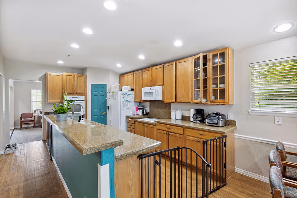 A bright senior facility kitchen with a long island, wooden cabinets, white appliances, a teal door, and a safety gate.