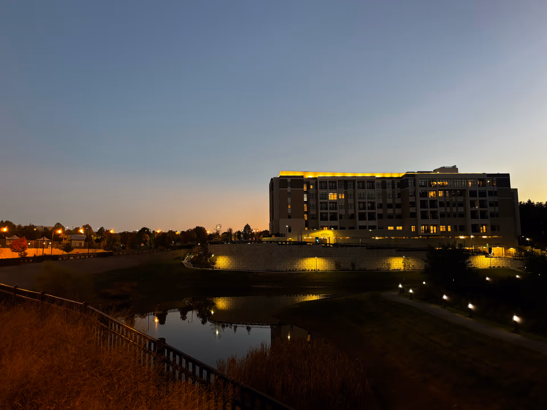Evening view of a large multi-story building illuminated with yellow lights along the roofline and windows. The building is situated on a raised area with a stone retaining wall and overlooks a small pond with reflections of the lights. There is a pathway with small lights leading towards the building, and trees and streetlights are visible in the background under a clear sky.