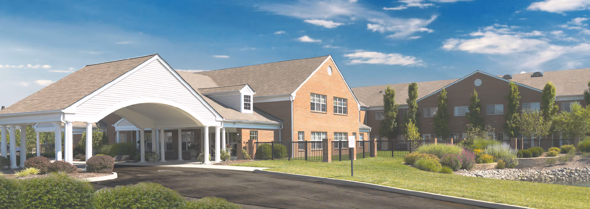 Exterior view of Villaspring Of Erlanger senior living facility showing a large brick building with multiple windows, a covered entrance with white columns, landscaped greenery, bushes, and a clear blue sky with some clouds.