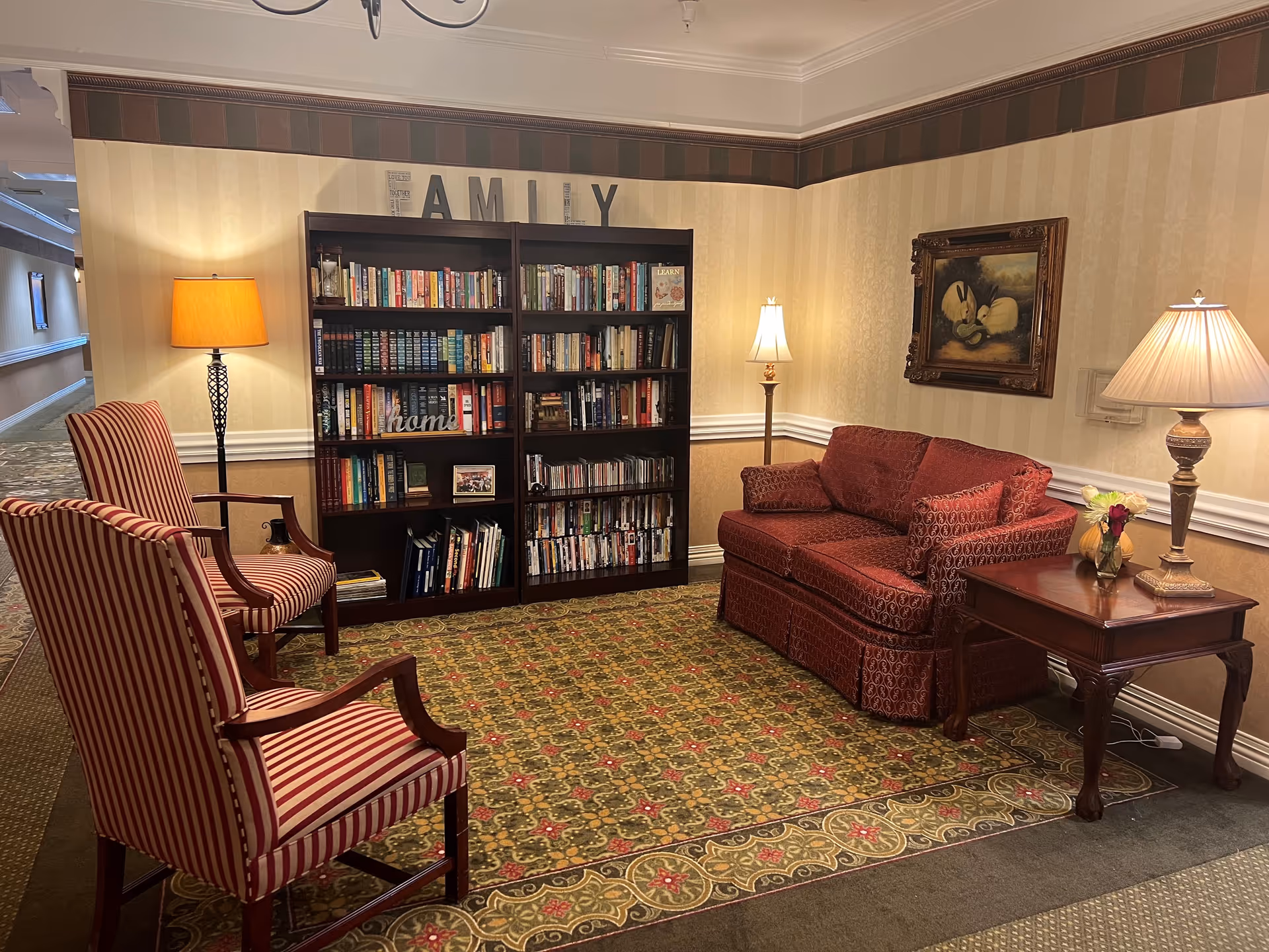 A cozy sitting area in a senior living facility with two striped armchairs, a red patterned loveseat, a wooden side table with a lamp and flowers, a bookshelf filled with books, and three lamps providing warm lighting. The wall behind the bookshelf has the word 'FAMILY' displayed, and there is a framed painting on the adjacent wall. The area is carpeted with a patterned rug.