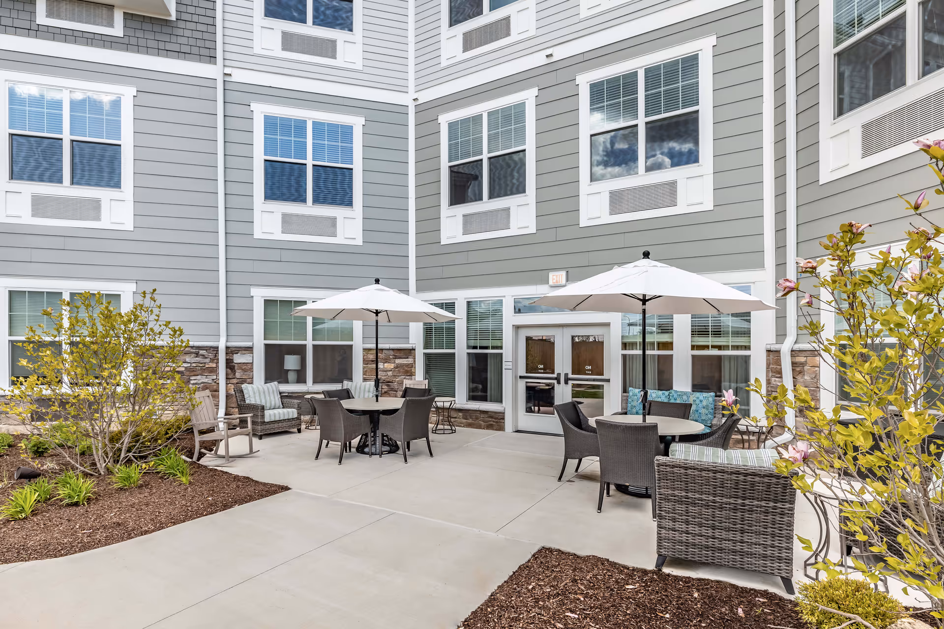 Outdoor patio area at StoryPoint Farmington Hills featuring round tables with umbrellas, wicker chairs, cushioned seating, and surrounding landscaping with bushes and flowers. The building exterior is gray with white trim and multiple windows.