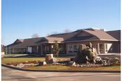 Exterior view of a single-story senior living facility building with a landscaped front yard featuring rocks and a small water fountain, under a clear blue sky.