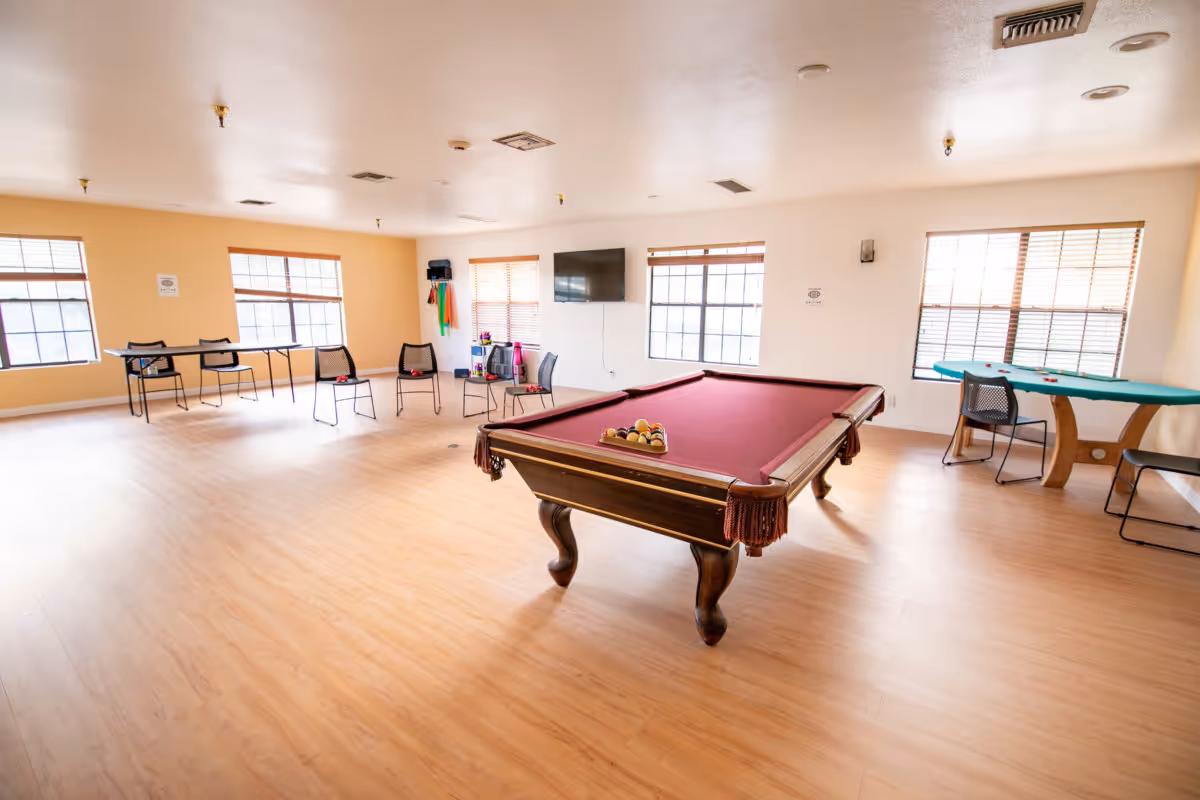 A spacious recreational room with a pool table in the center, several chairs arranged around the room, a ping pong table on the left side, and a card table on the right side near windows with blinds. The room has light wood flooring and white walls with large windows letting in natural light.
