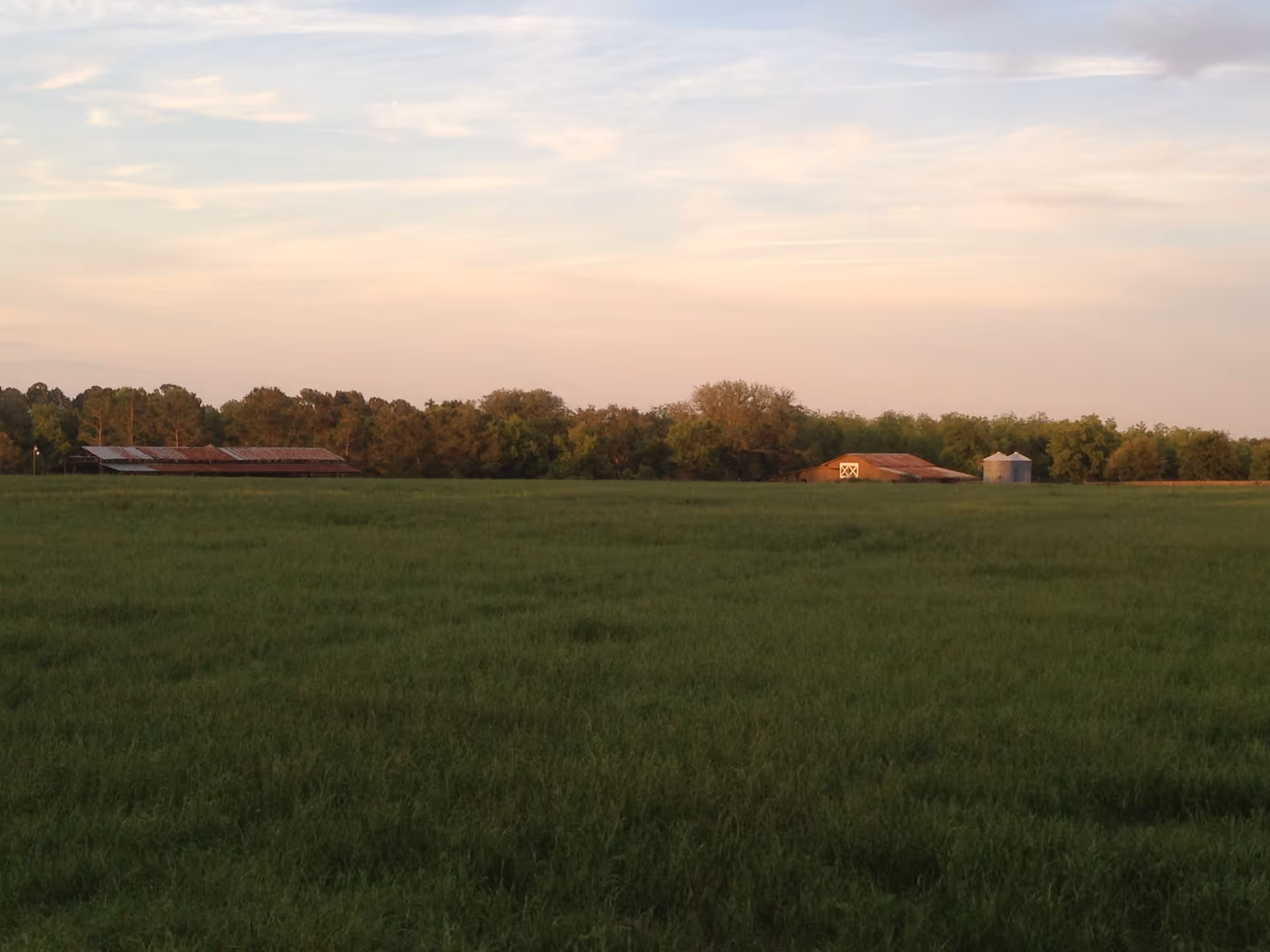 A wide open green field with two rustic barns and two silos in the distance under a partly cloudy sky during sunset.