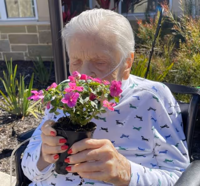 An elderly person sitting outdoors in a wheelchair, holding and smelling a small potted plant with bright pink flowers. The person is wearing a white shirt with a colorful dog pattern and has an oxygen tube in their nose. There are plants and a building visible in the background.