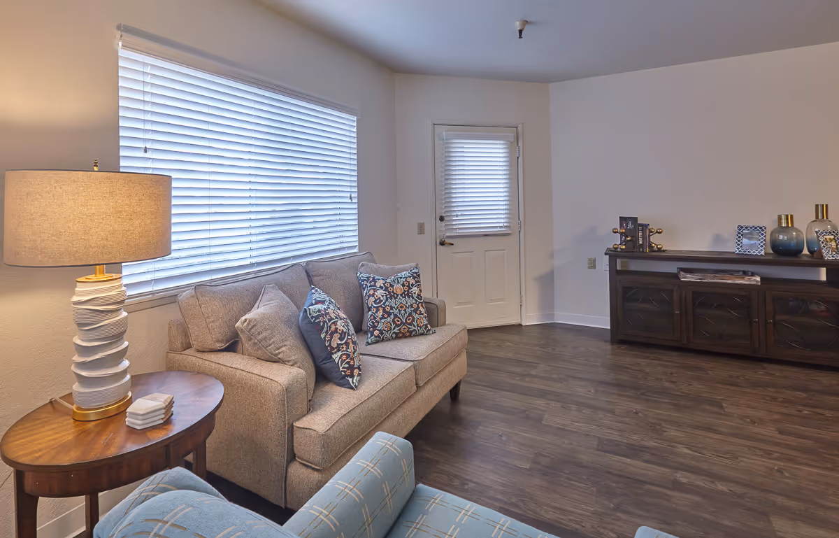 A cozy living room with a beige sofa adorned with patterned and solid pillows, a wooden side table with a modern white lamp, a blue armchair with a subtle pattern, a large window with white blinds, a white door with a window also covered by blinds, and a dark wooden cabinet decorated with vases, picture frames, and books on a wooden floor.