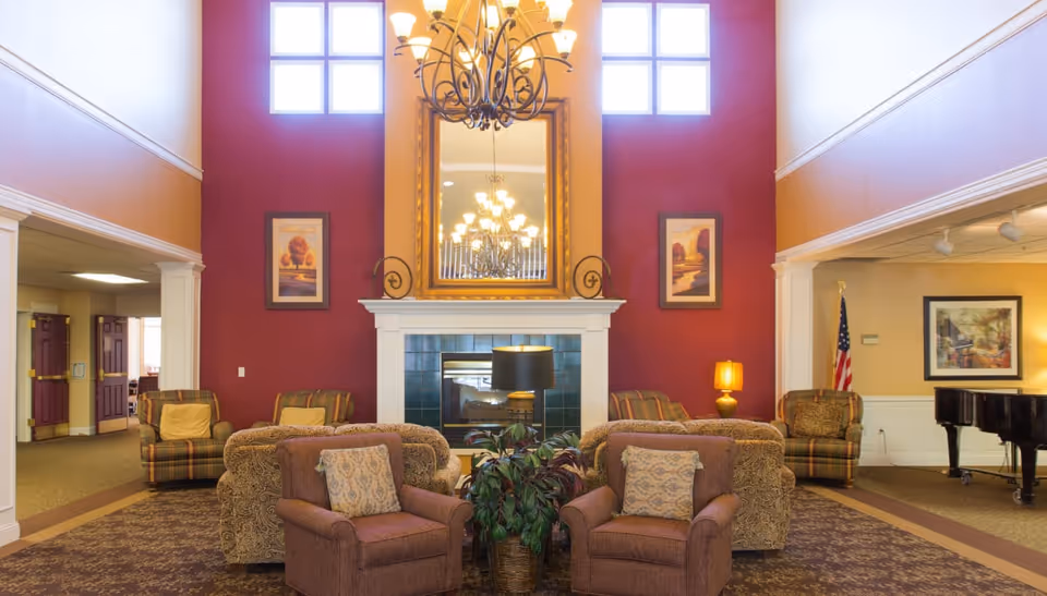 A spacious living room area with a high ceiling and large windows near the top. The room features a fireplace with a large ornate mirror above it, flanked by two framed paintings on a red accent wall. There are multiple upholstered armchairs and sofas arranged around a central coffee table with a plant. A chandelier hangs from the ceiling, and there is a piano and an American flag in the background.