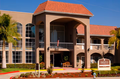 Front facade of a two-story beige senior living building with an arched entrance, red tile roof, balconies, palm trees, and an Emeritus sign.