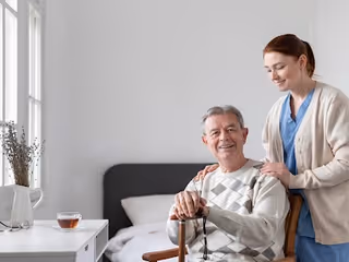 An elderly man sitting on a bed holding a cane, smiling, with a woman standing behind him gently resting her hands on his shoulders in a bright, simple bedroom setting.