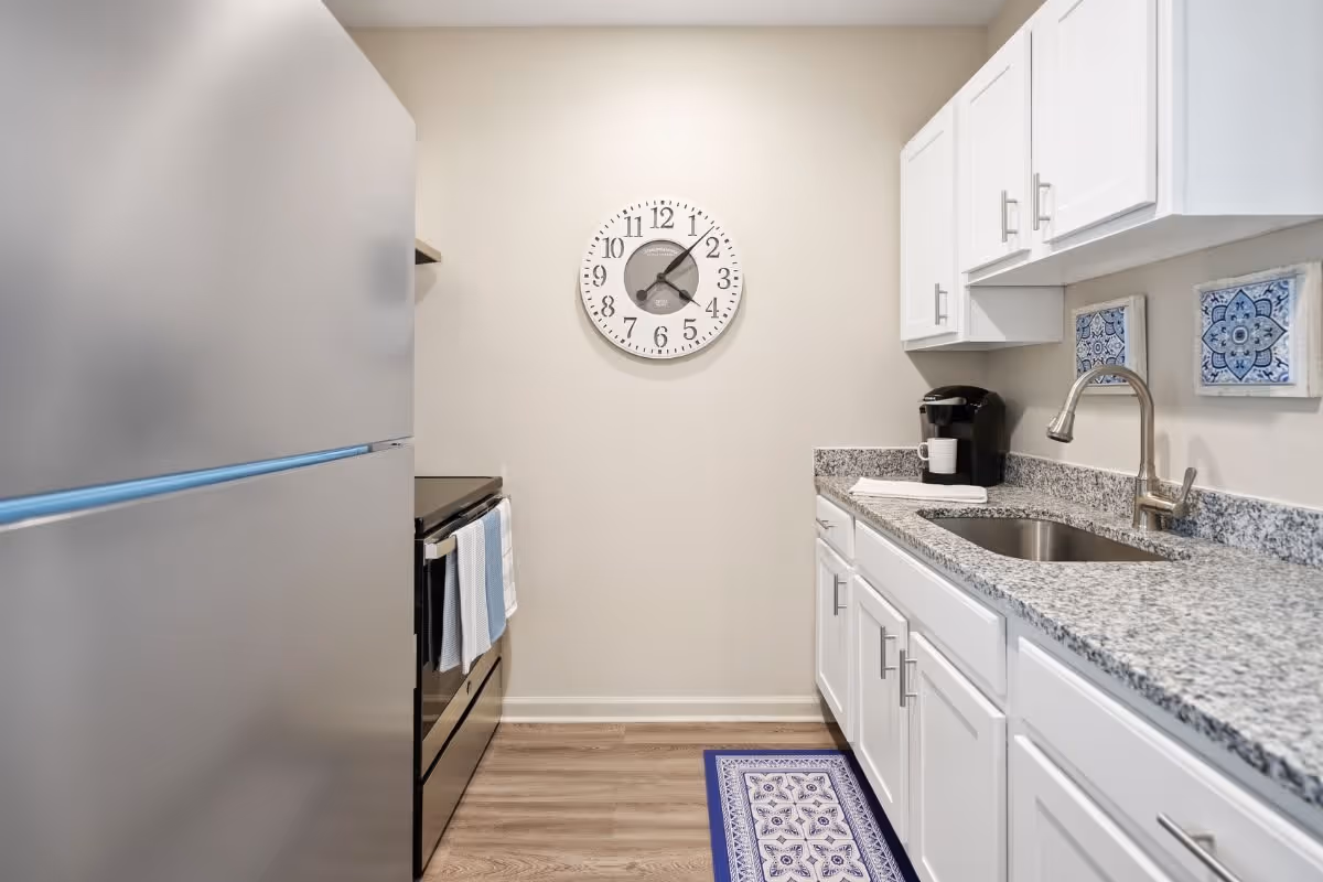 A modern kitchen with white cabinets, granite countertops, a stainless steel refrigerator, a black stove with a blue and white towel hanging on the handle, a stainless steel sink with a faucet, a coffee maker, two decorative blue and white tiles on the wall, a blue patterned rug on the wooden floor, and a round wall clock showing the time as 7:10.