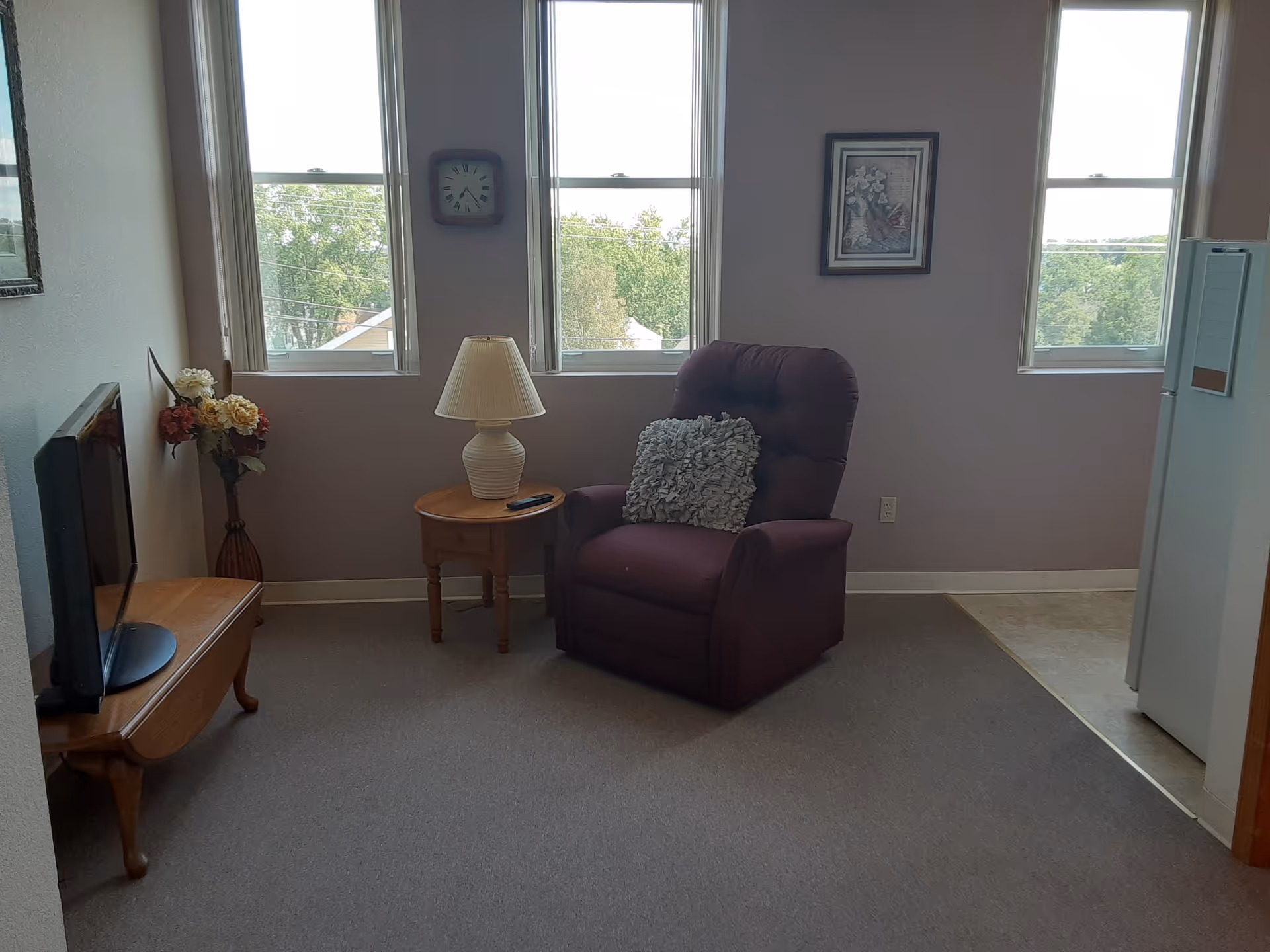 A cozy living room area with a purple recliner chair featuring a decorative pillow, a wooden side table with a lamp and remote control, a wooden TV stand with a flat-screen TV, a vase with artificial flowers, three large windows letting in natural light, a wall clock, and a framed picture on the wall.