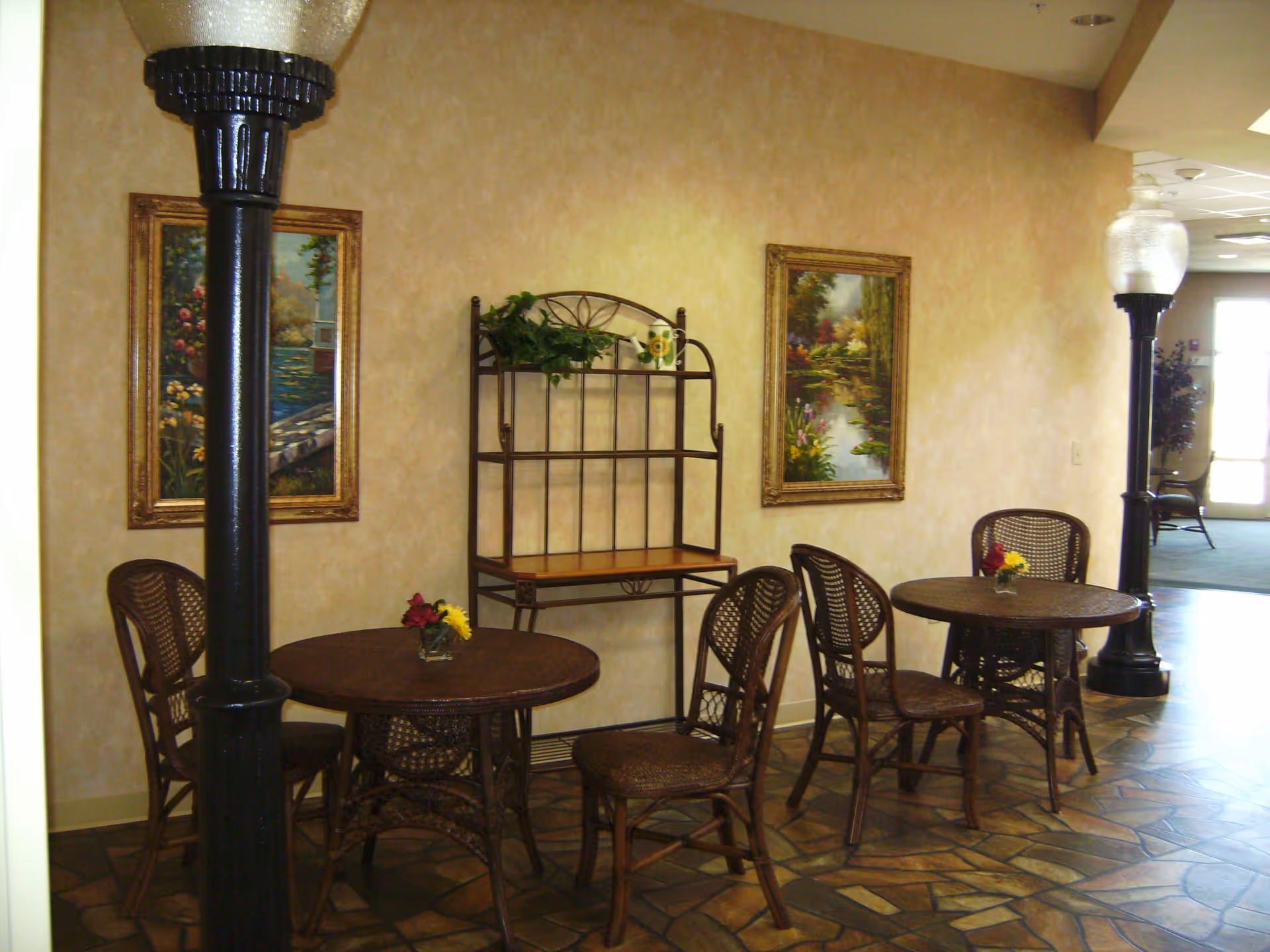 Interior seating area with two round wicker tables, each surrounded by wicker chairs. Each table has a small vase with flowers. There are two tall black lamp posts with frosted glass tops, two framed landscape paintings on the beige wall, and a metal shelf unit with a plant and decorative items. The floor has a patterned tile design.