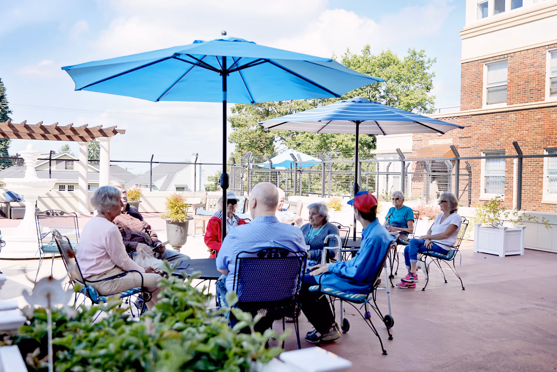 A group of elderly people sitting and socializing outdoors under blue umbrellas on a patio with metal chairs and tables. The setting includes a brick building, potted plants, and a clear sky.