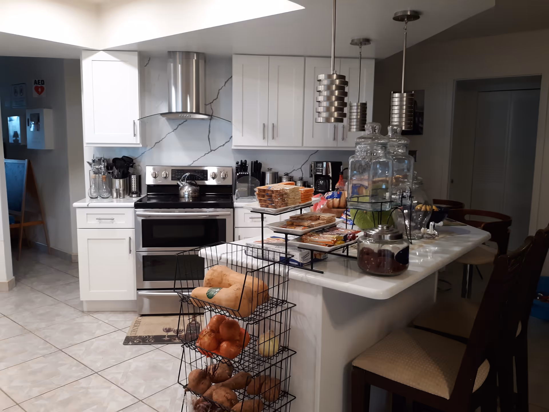 Modern communal kitchen with white cabinets and a stainless-steel stove, an island counter lined with snacks and glass dispensers, and bar stools.