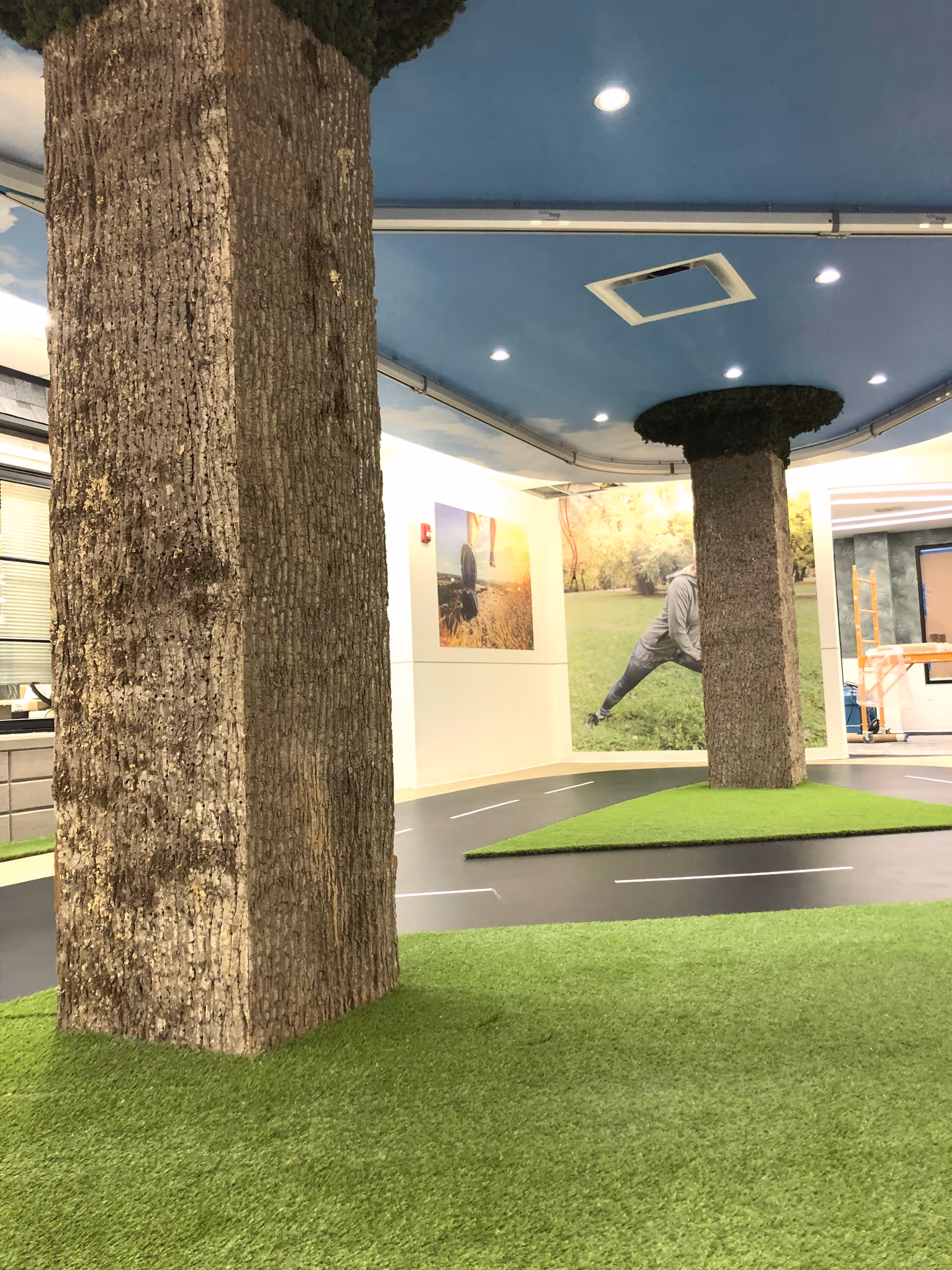 Indoor space with artificial grass flooring and two large columns designed to look like tree trunks with foliage at the top. The ceiling is painted blue with recessed lighting and an air vent. The walls have large photos depicting outdoor scenes with people exercising and enjoying nature.