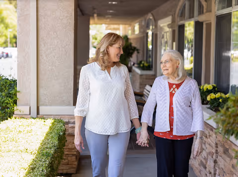 An elderly woman and a younger woman holding hands and walking together along a covered outdoor walkway next to a building with windows and flower pots.