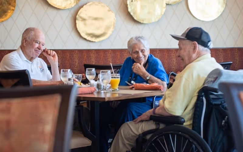 Three elderly people sitting around a dining table in a restaurant or dining area, smiling and engaging in conversation. One person is in a wheelchair, and there are glasses of water and orange juice on the table. Decorative round gold plates hang on the wall behind them.