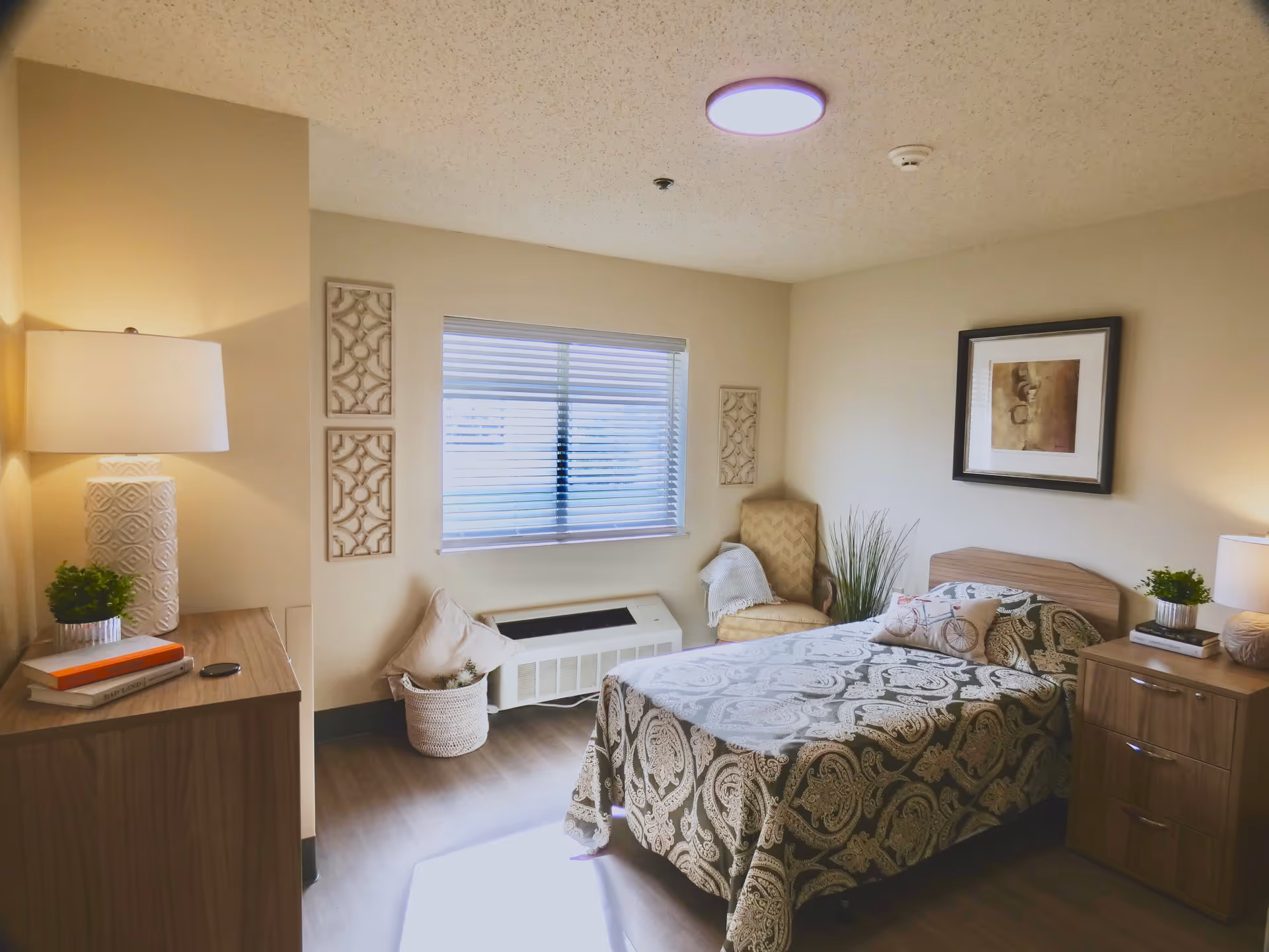 A cozy bedroom in a senior living facility with a single bed covered in a patterned bedspread, a wooden nightstand with a lamp and plant, a wooden dresser with a lamp and books, a cushioned armchair with a throw blanket, decorative wall art, and a window with blinds letting in natural light.