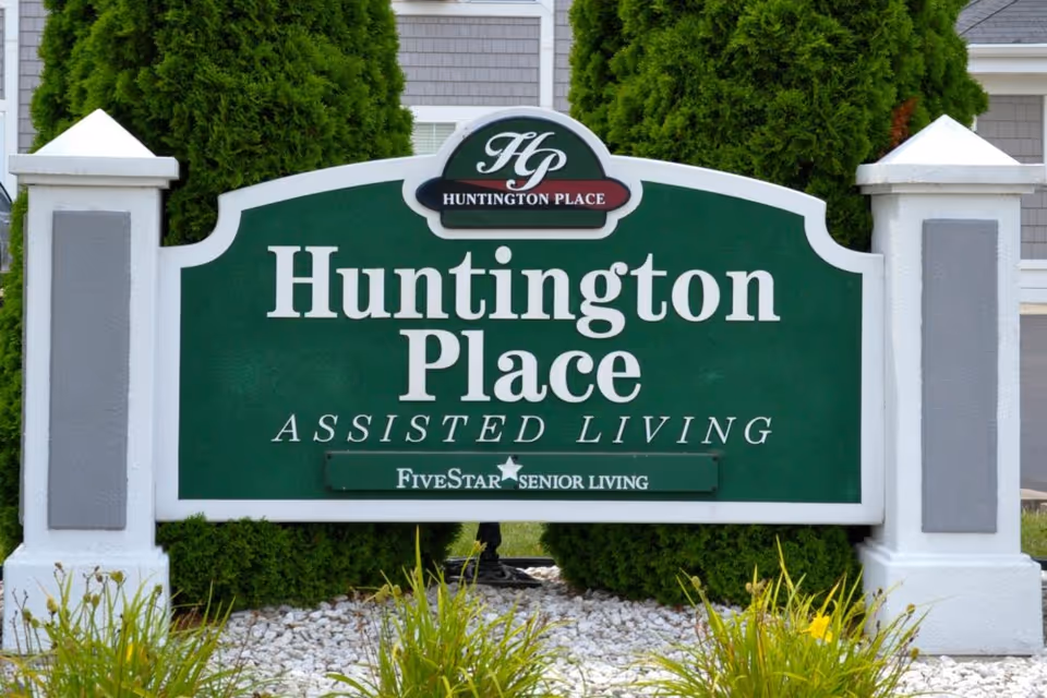 Green and white sign for Huntington Place Assisted Living, with the logo HP and the text Five Star Senior Living, surrounded by greenery and landscaping.