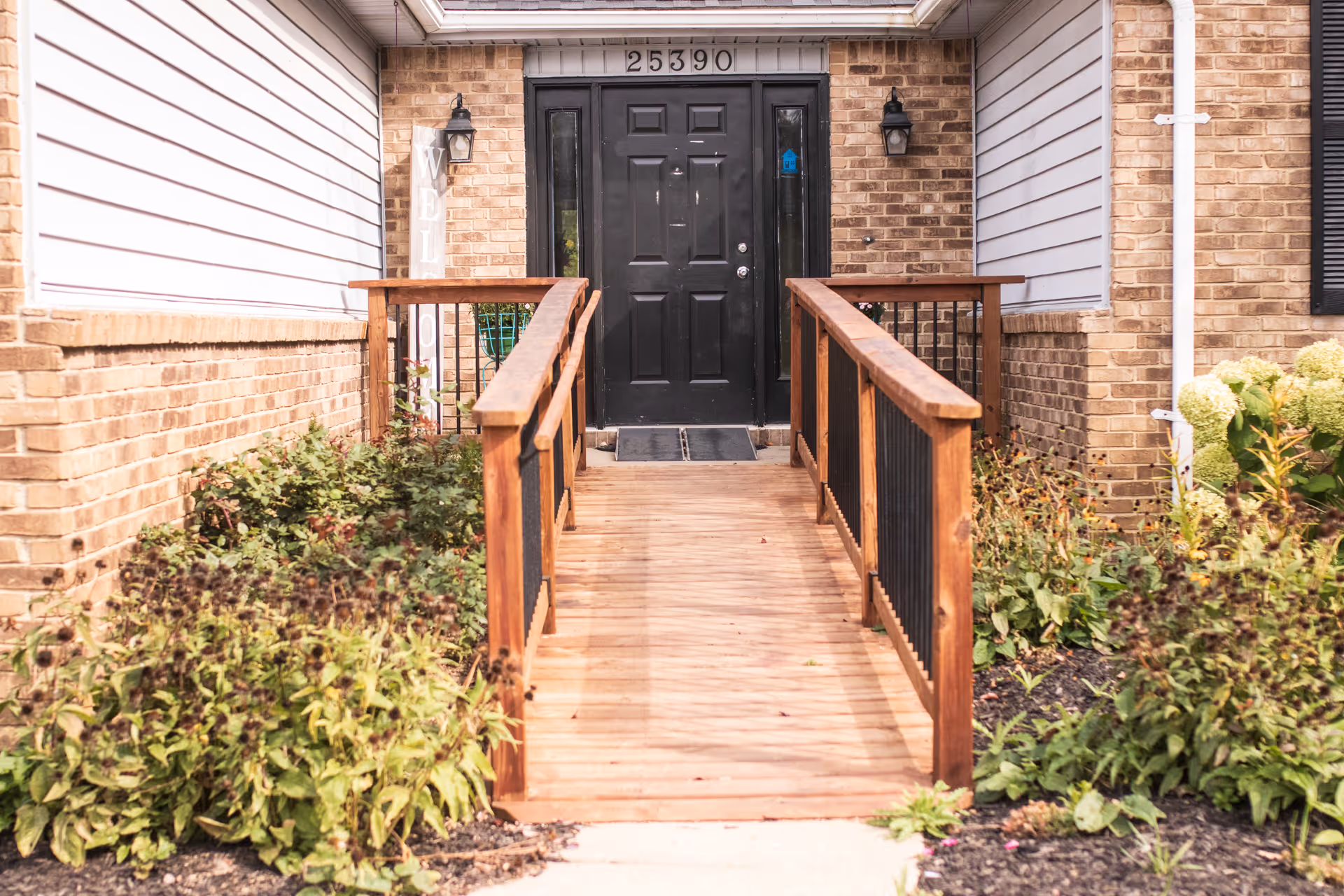 Front entrance of a building with a wooden ramp leading to a black door. The building has brick walls and white siding, with plants and shrubs on either side of the ramp. The address number 25390 is displayed above the door, and there are two wall-mounted lantern lights on either side of the door.