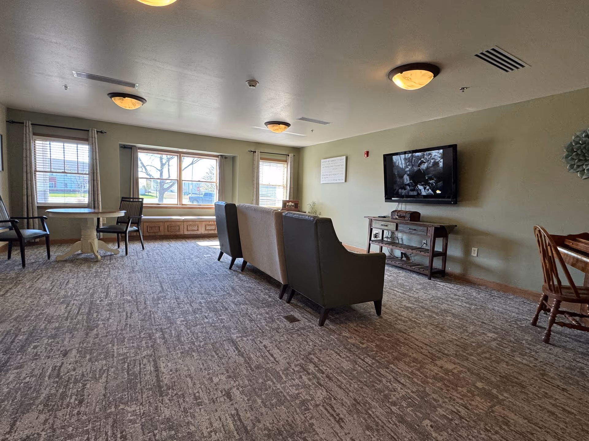 A spacious living room area with a large window letting in natural light. The room features a carpeted floor, two armchairs and a sofa facing a wall-mounted television. There is a small wooden table with a chair on the right side and a round table with chairs near the windows. The walls are painted light green and decorated with a wall sign and a decorative piece.